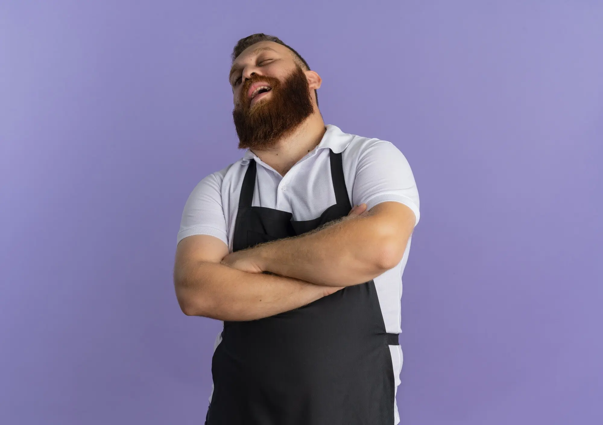 Genervter, professioneller, bärtiger, männlicher Barbier in Schürze mit vor der Brust verschränkten Armen steht vor einer violetten Wand.