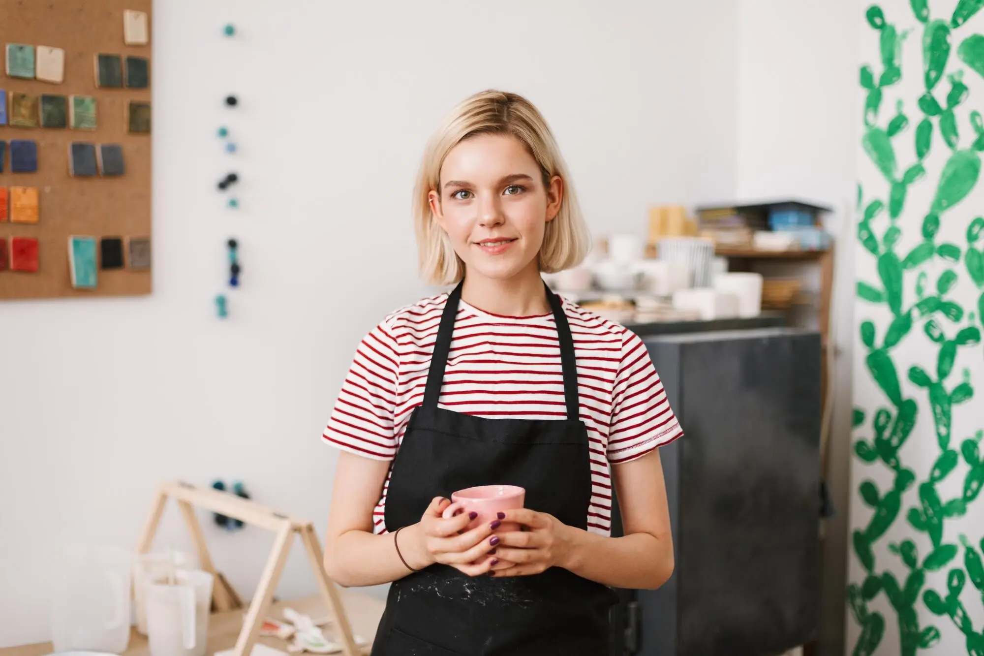 In einer Töpferwerkstatt hält ein hübsches Mädchen in schwarzer Schürze eine handgefertigte Tasse in den Händen und blickt verträumt in die Kamera.