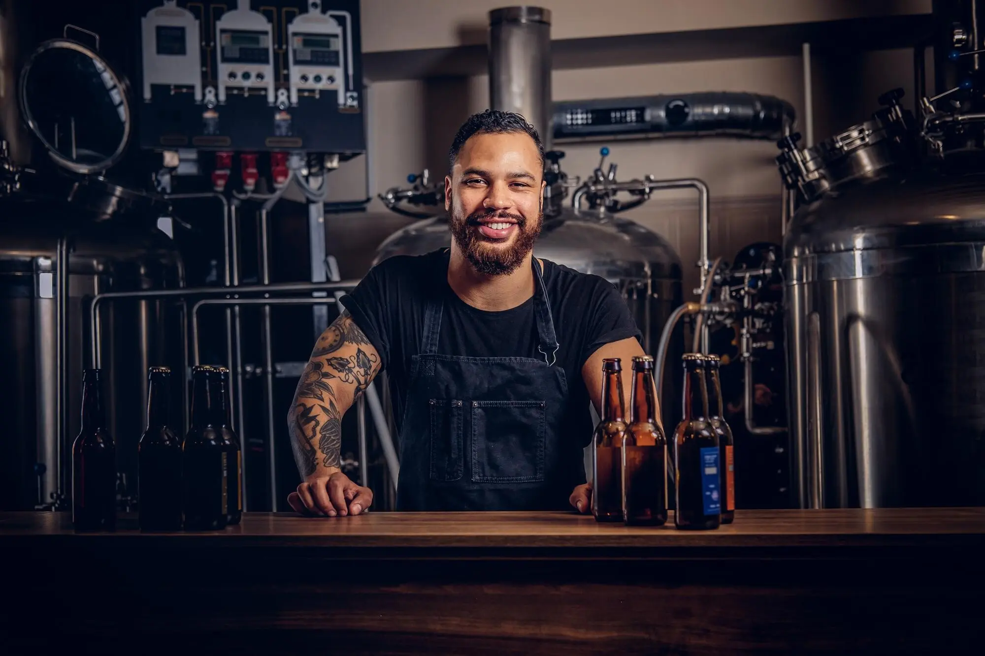 Porträt eines stilvollen, bärtigen, dunkelhäutigen Mannes mit einem Tattoo auf der Hand, der hinter der Theke in einer Brauerei steht.